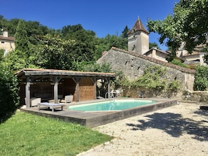 Ein Schwimmbecken mit Holzterrasse, ein Steinhaus mit Turm und umgebende Bepflanzung.