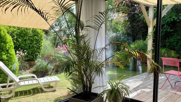 Une pièce dotée d’une grande fenêtre, d’une plante en pot, d’une chaise blanche et d’une terrasse en bois visible à travers la vitre.