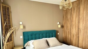 Une chambre avec une tête de lit de couleur turquoise, un lit recouvert de draps blancs, une armoire en bois et une petite table de chevet ronde avec une lampe.