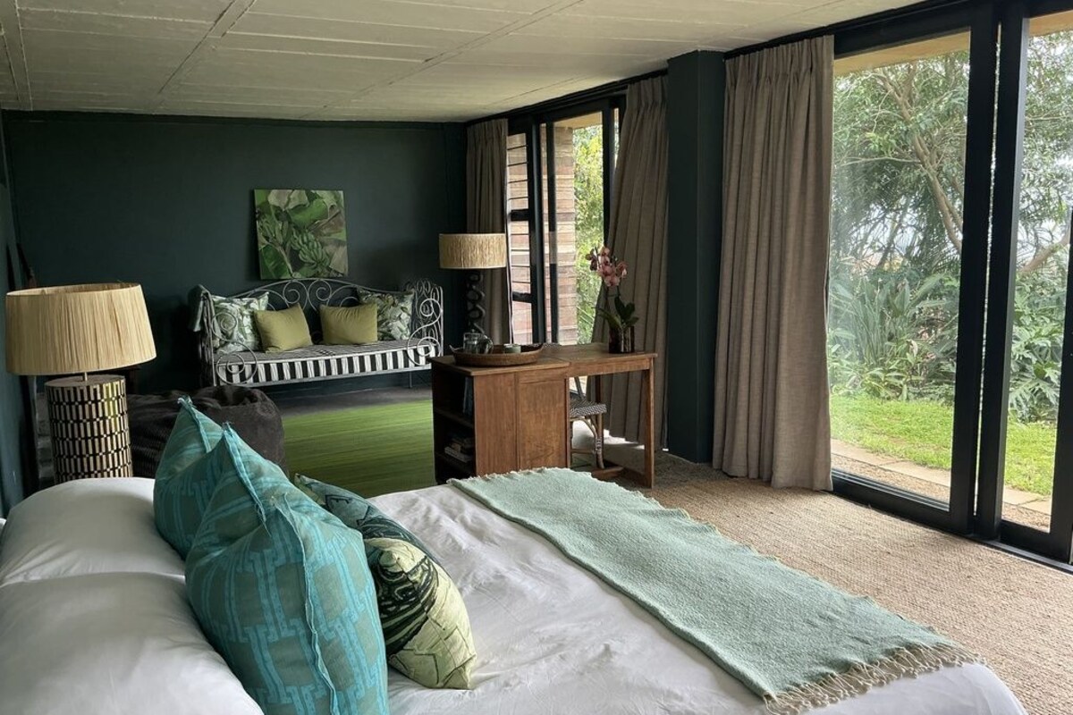 Un dormitorio con cama, lámparas de noche, un escritorio y vistas a zonas verdes a través de amplios ventanales.