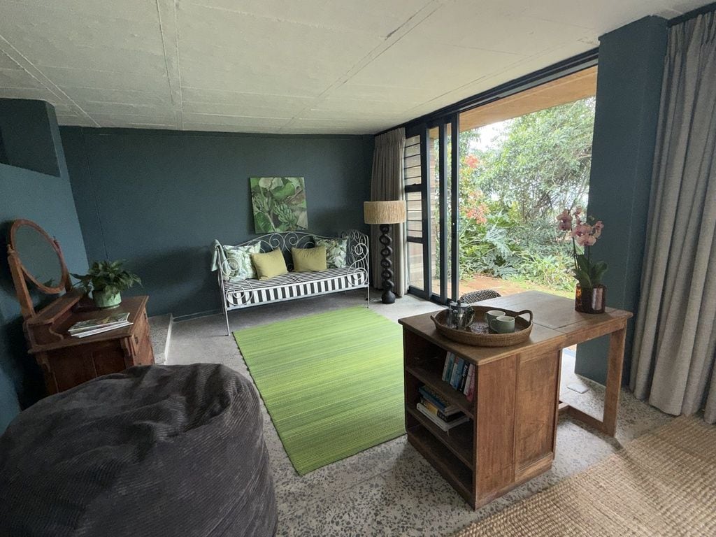 Une pièce avec un tapis vert, une table d’appoint en bois, un banc avec des coussins, une bibliothèque et une vue sur la verdure à l’extérieur.