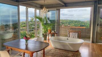 A modern bathroom with a freestanding bathtub, a wooden table with chairs, and a large window offering a view of a hilly landscape.