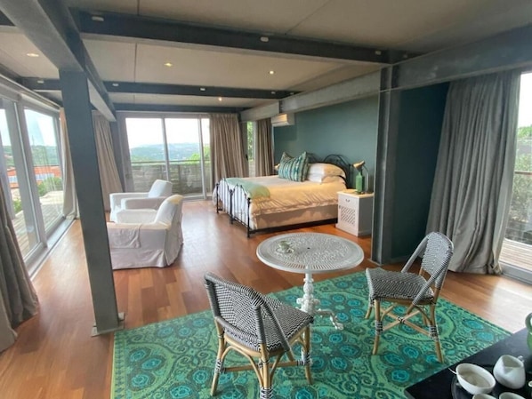 A hotel room with a bed, a sitting area with a table and chairs, and a view through large windows.