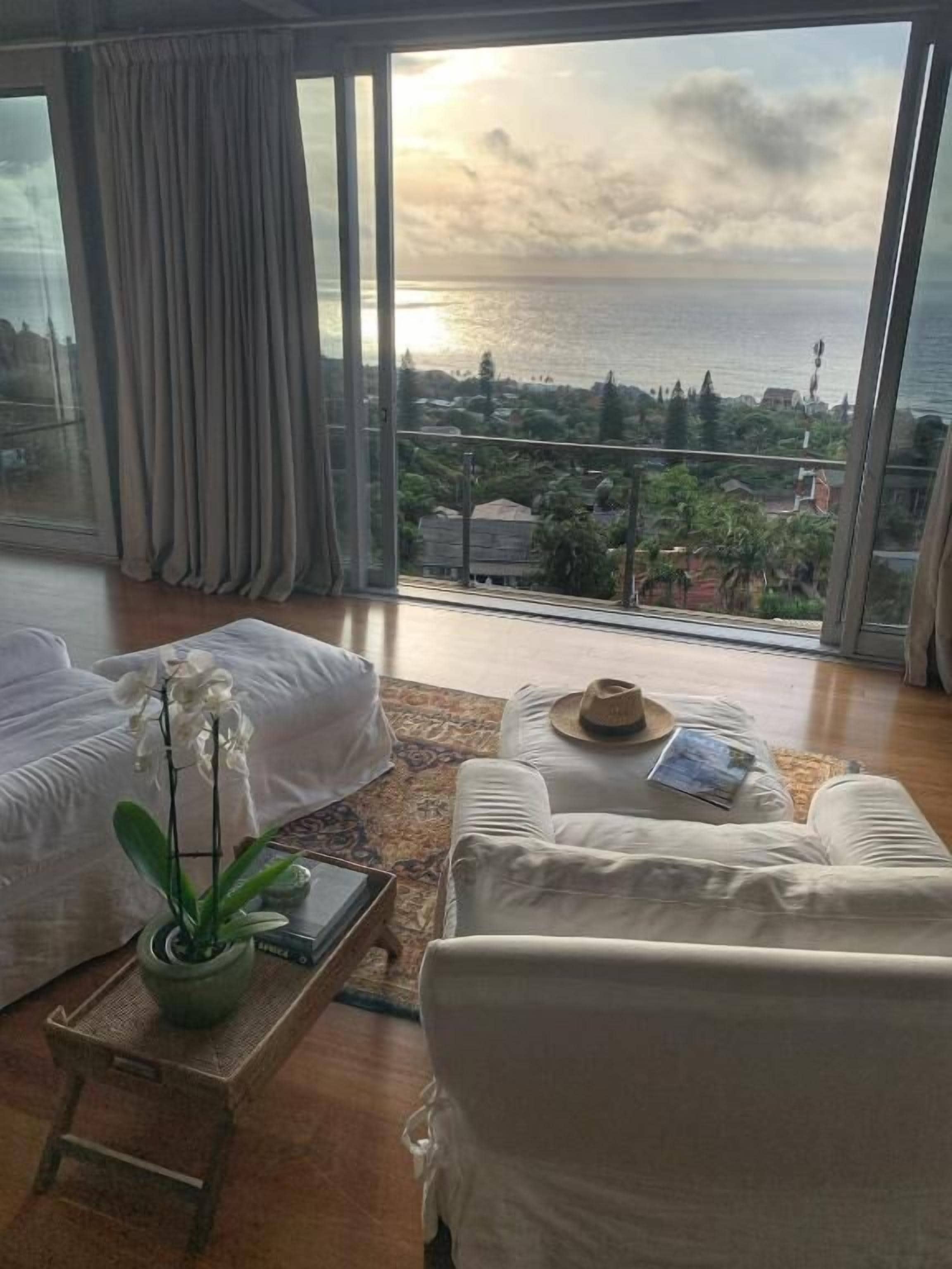 A room with a large window overlooking a body of water, a sofa, a coffee table with a hat and book, and a potted orchid.