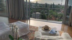 A room with a large window overlooking a body of water, a sofa, a coffee table with a hat and book, and a potted orchid. - The Seaglass (Durban)