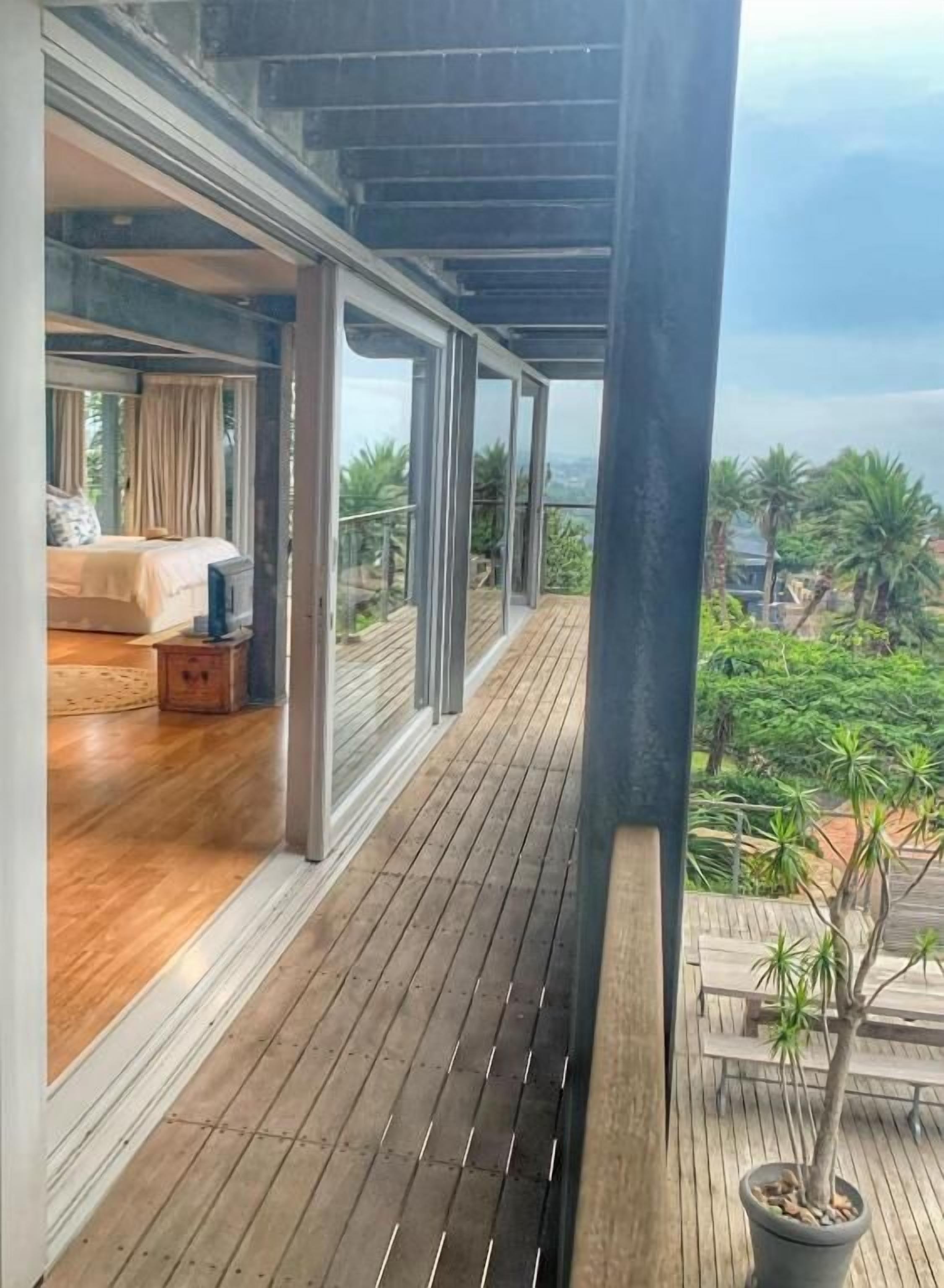 A balcony with a wooden floor, a bed with a white headboard, and a view of greenery.