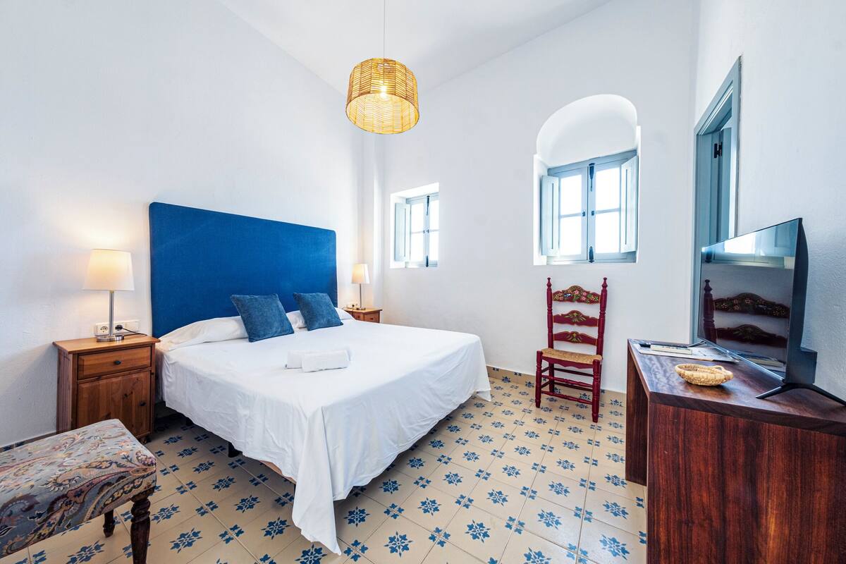 A bedroom with a large bed, a wooden nightstand, a chair, a desk, a television, and a hanging light fixture.