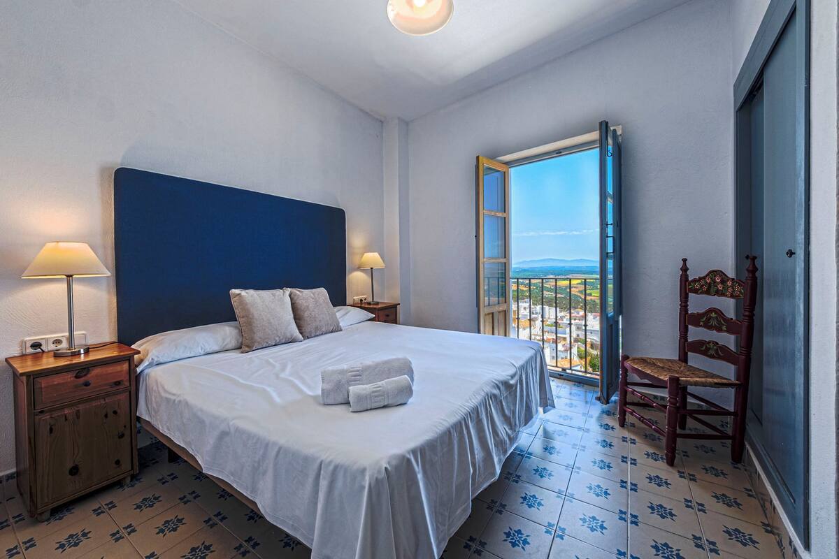 A hotel room with a bed, two bedside lamps, a wooden nightstand, a chair, and a balcony with a view.
