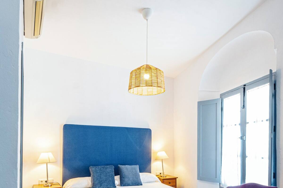 A bedroom with a large bed, two bedside tables, a hanging light fixture, and a window with shutters.