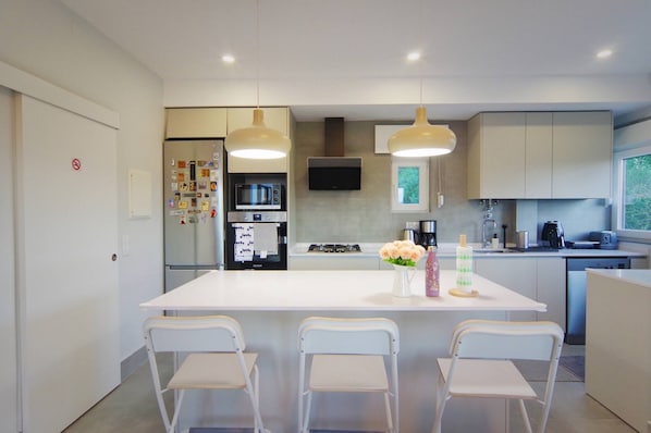 A modern kitchen with a central island, white chairs, a flat-screen TV, and a window overlooking greenery. - Entire Cascais New Mordern Apartment-3 Bedrooms (Cascais)