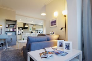 A modern living room with a grey sofa, a white coffee table with magazines, a bookshelf, a kitchen area, and a framed picture on the wall. - Entire Cascais New Mordern Apartment-3 Bedrooms (Cascais)