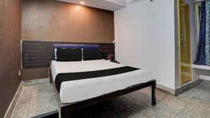 Una habitación de hotel con una cama, cabecera de madera, una mesita de noche pequeña y una puerta que da a un baño.