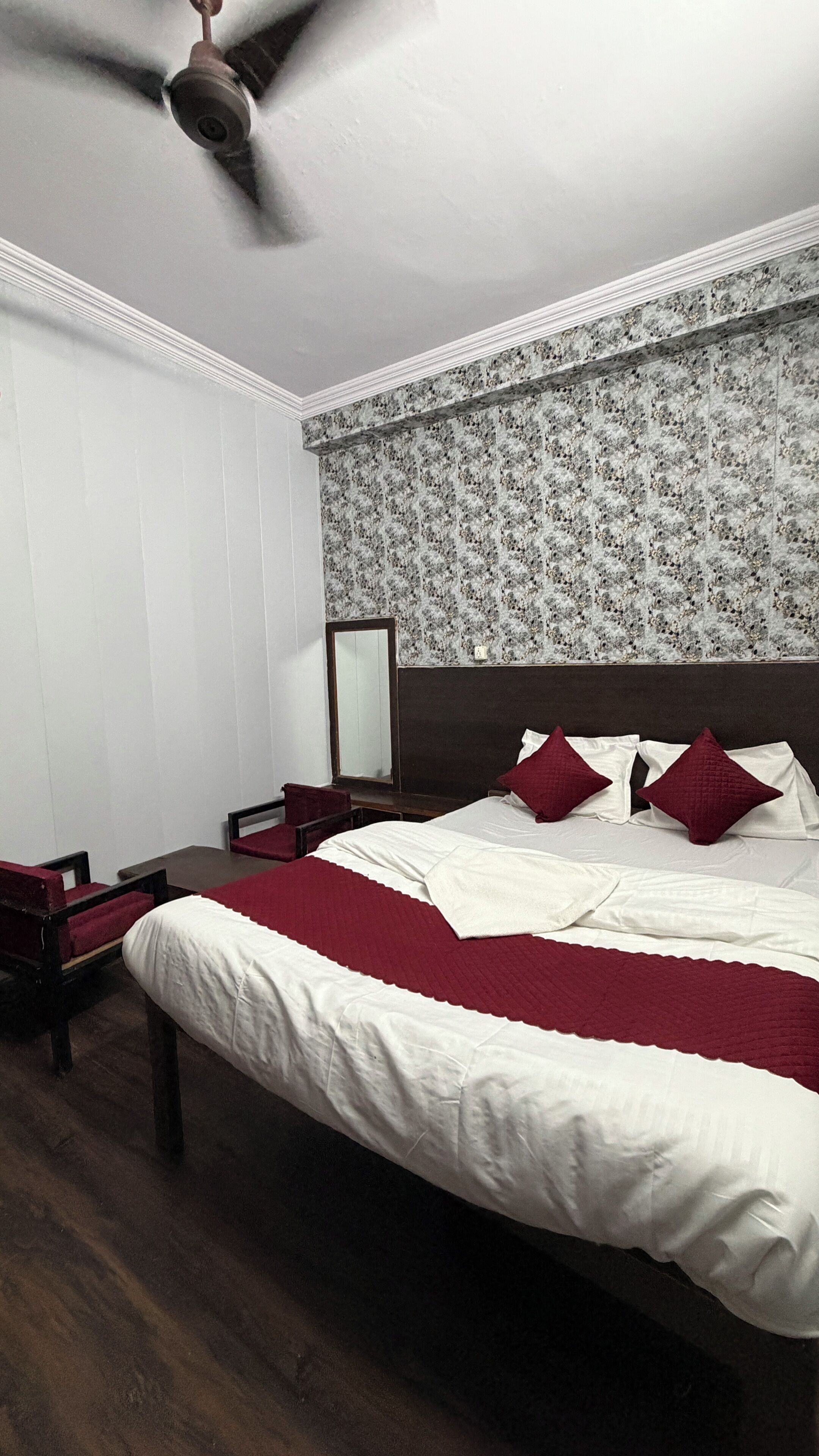 A bedroom with a large bed, a ceiling fan, a wooden nightstand, a mirror, and a chair.