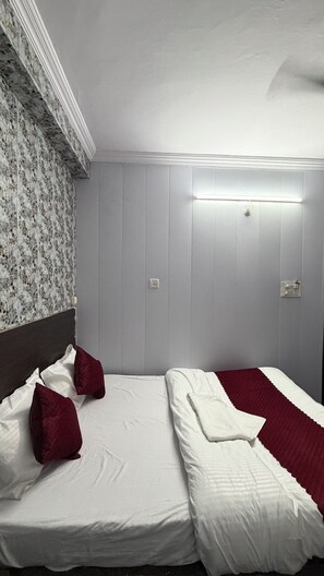 A bedroom with a bed, two pillows, a bedside table, a wall-mounted light, and a patterned wallpaper. - Hotel The Royal Palace (Lucknow)