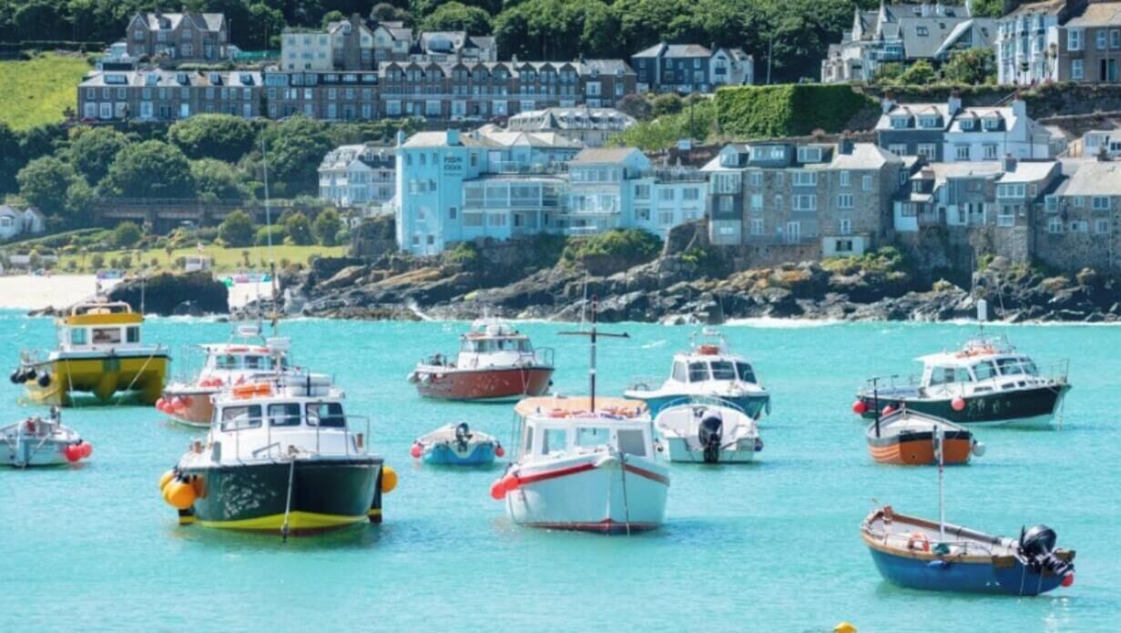 A coastal town with numerous boats docked in the water, buildings with light blue facades, and a rocky shoreline.