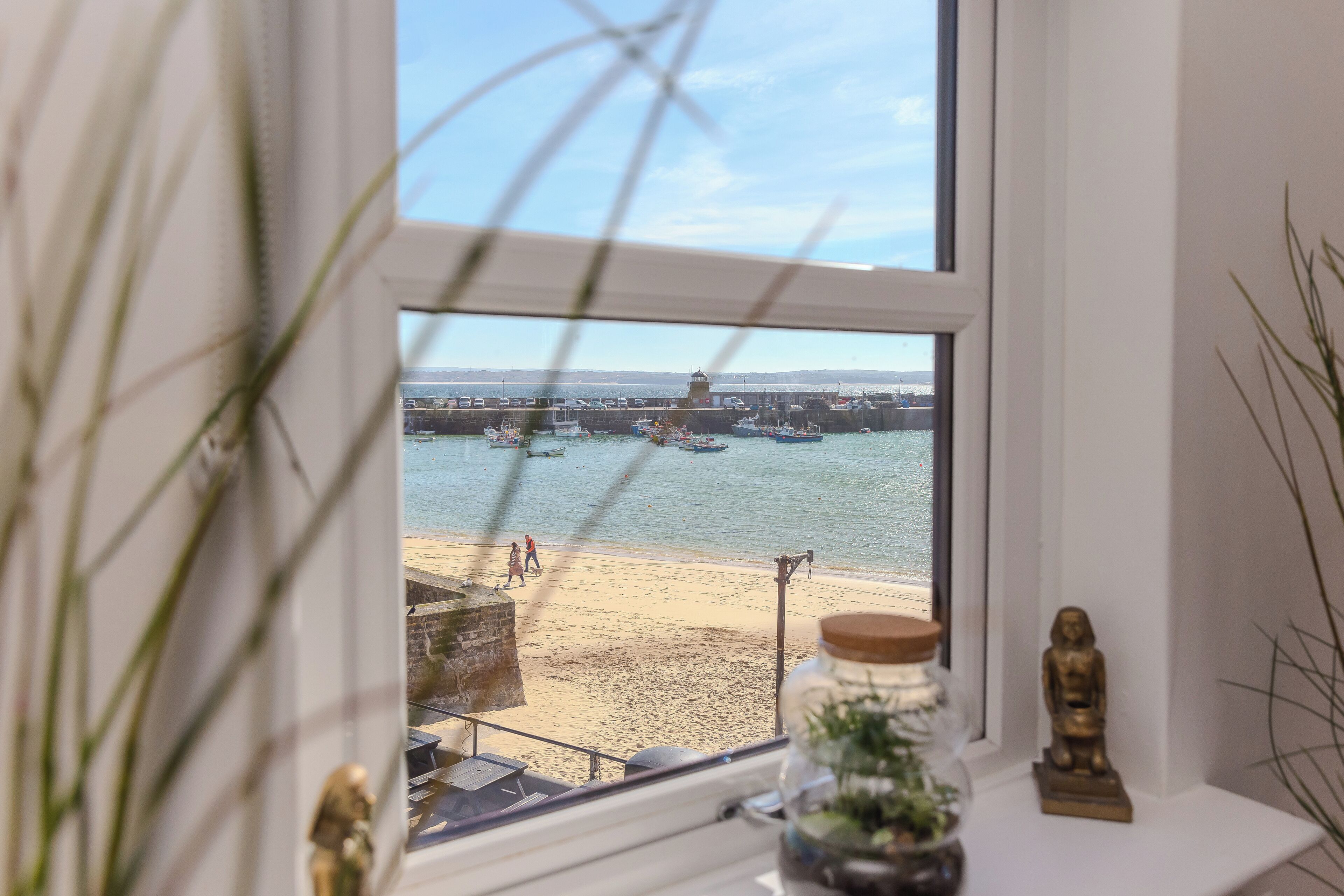 A view through a window of a beach with boats, a statue, and a jar with a plant.