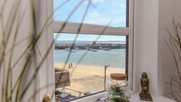 A view through a window of a beach with boats, a statue, and a jar with a plant.