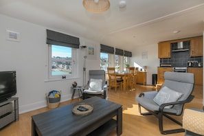 Ein modernes Wohnzimmer mit Fernseher, ein Essbereich mit Tisch und Stühlen, eine Küche mit Holzschränken und Blick auf die Stadt.