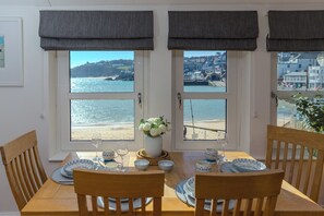A dining table set with plates, bowls, and wine glasses, with a view of the beach and buildings through large windows. - Harts Loft | On the Beach | Panoramic Sea Views | By Harbour | Pet Friendly (St Ives)