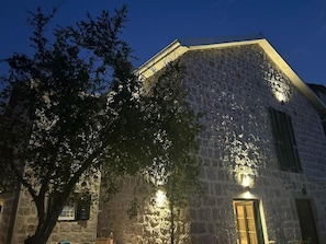 A stone building with a sloped roof, illuminated by exterior lights, and a tree in the foreground.