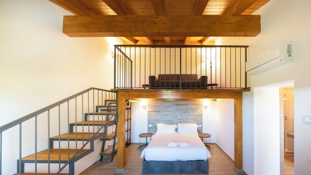 Ein Schlafzimmer mit Bett, Treppe, einer Couch auf einer erhöhten Ebene und einer Holzbalkendecke.