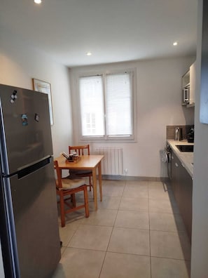 Una cocina compacta con refrigerador, mesa de comedor con sillas, ventana con persianas y microondas.