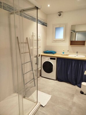 A bathroom with a glass shower enclosure, a washing machine, a sink, and a mirror.