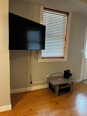 Une pièce dotée d’un téléviseur à écran plat fixé au mur, d’une fenêtre avec des stores, d’une petite table en bois sur laquelle se trouve un appareil noir, et d’un plancher en bois.