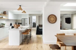 Eine moderne Küche mit weißen Schränken, einer zentralen Kochinsel mit Sitzgelegenheiten, einem Essbereich mit Holztisch und einer Treppe zu einer weiteren Ebene.