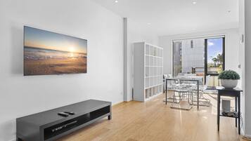 Una sala de estar moderna con un televisor de pantalla plana, un mueble de entretenimiento negro, una zona de comedor con una mesa y sillas de vidrio, y un ventanal con vista a la ciudad.