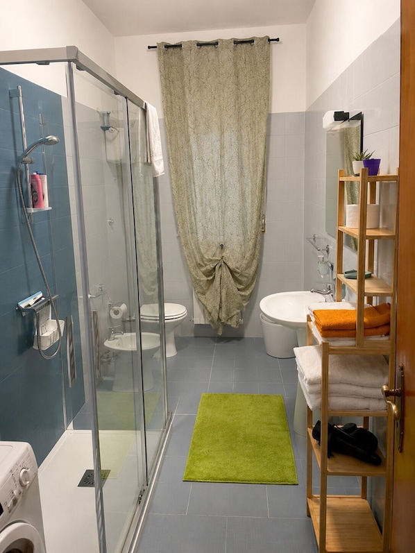 A bathroom with a glass shower enclosure, a toilet, a sink, and a wooden shelf with towels. - Cozy Rooms KLM (Santeramo In Colle)