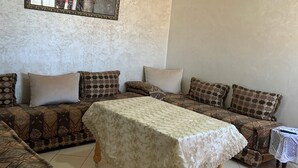 Een woonkamer met een zithoek, een tafel met kantkleed, een kroonluchter en een kunstwerk in een lijst aan de muur.