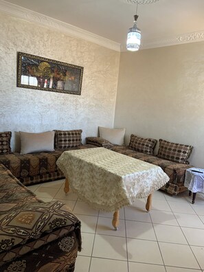 Un salon comprenant un ensemble de canapés, une table recouverte d’une nappe en dentelle, un lustre et une œuvre d’art encadrée accrochée au mur.