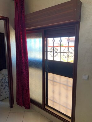 Une pièce dotée d’une porte-fenêtre coulissante, d’un rideau à motifs, d’un lit recouvert d’une couvre-lit à motifs et d’un sol carrelé.