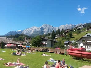 Eine grüne Außengegend mit sonnenden Personen, einem roten Boot und Häusern mit Blick auf die Berge im Hintergrund.