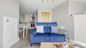 En moderne stue med en blå sofa, et træbord med en bakke druer og kaffekopper, og et køkken i baggrunden.