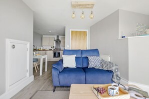 A modern living room with a blue sofa, a wooden coffee table with a tray of grapes and coffee cups, and a kitchen in the background. - Marina View Suite, Astor House - Sea view, 250 meters from beach (Torquay)
