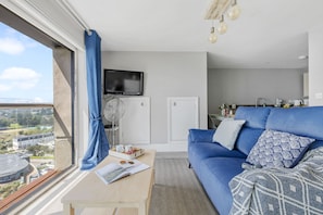 A modern living room with a blue sofa, a flat-screen TV, a fan, and a view of the city.