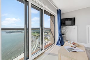 A room with large windows offering a city view, a flat-screen TV mounted on the wall, and a table with a book and a tray of items.