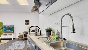 Una cocina moderna con un lavamanos de acero inoxidable, un horno empotrado y un microondas. Hay una planta en maceta sobre la encimera y una pequeña mesa con una silla.