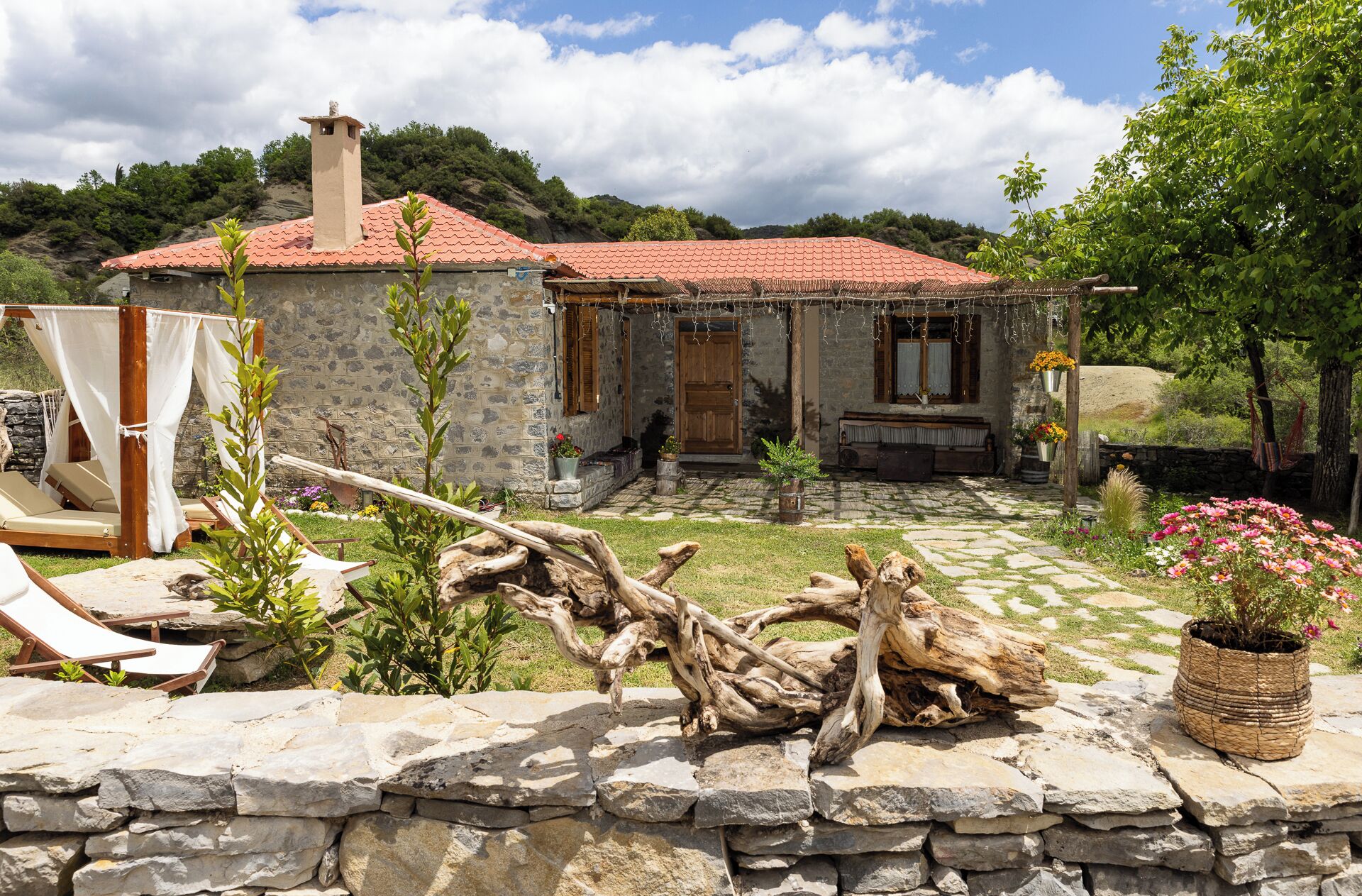 A stone house with a red-tiled roof, a stone wall, and a garden with a log bench and a canopy.