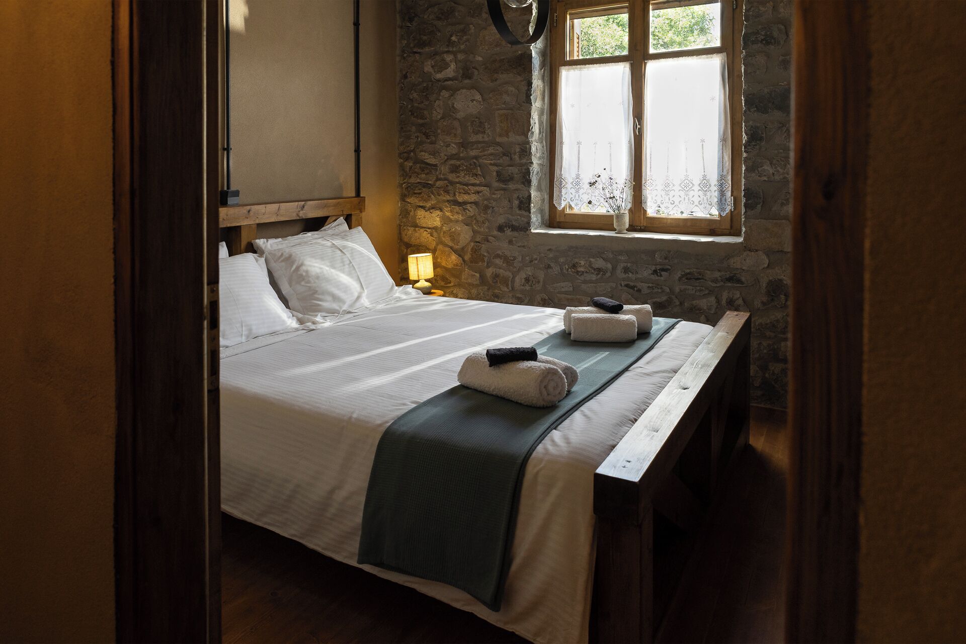Een bed met witte lakens, een houten hoofdbord, een raam met kantgordijnen en een stenen muur.