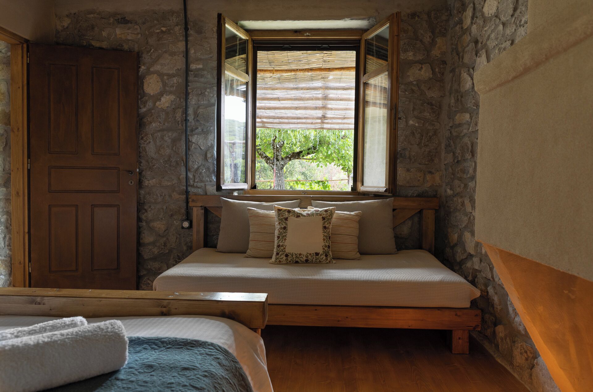 A bedroom with a stone wall, a wooden bed, a window with a view of greenery, and a door.