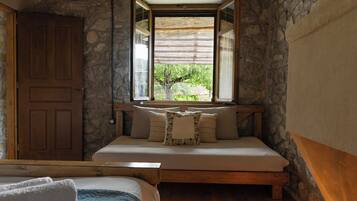 A bedroom with a stone wall, a wooden bed, a window with a view of greenery, and a door.