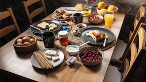 Une table de salle à manger en bois, sur laquelle sont disposés divers aliments pour le petit-déjeuner, une fenêtre donnant sur un paysage verdoyant et des murs en pierre.