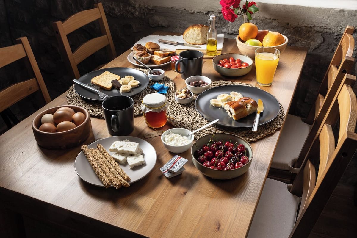 A wooden dining table with various breakfast items, a window with a view of greenery, and stone walls.