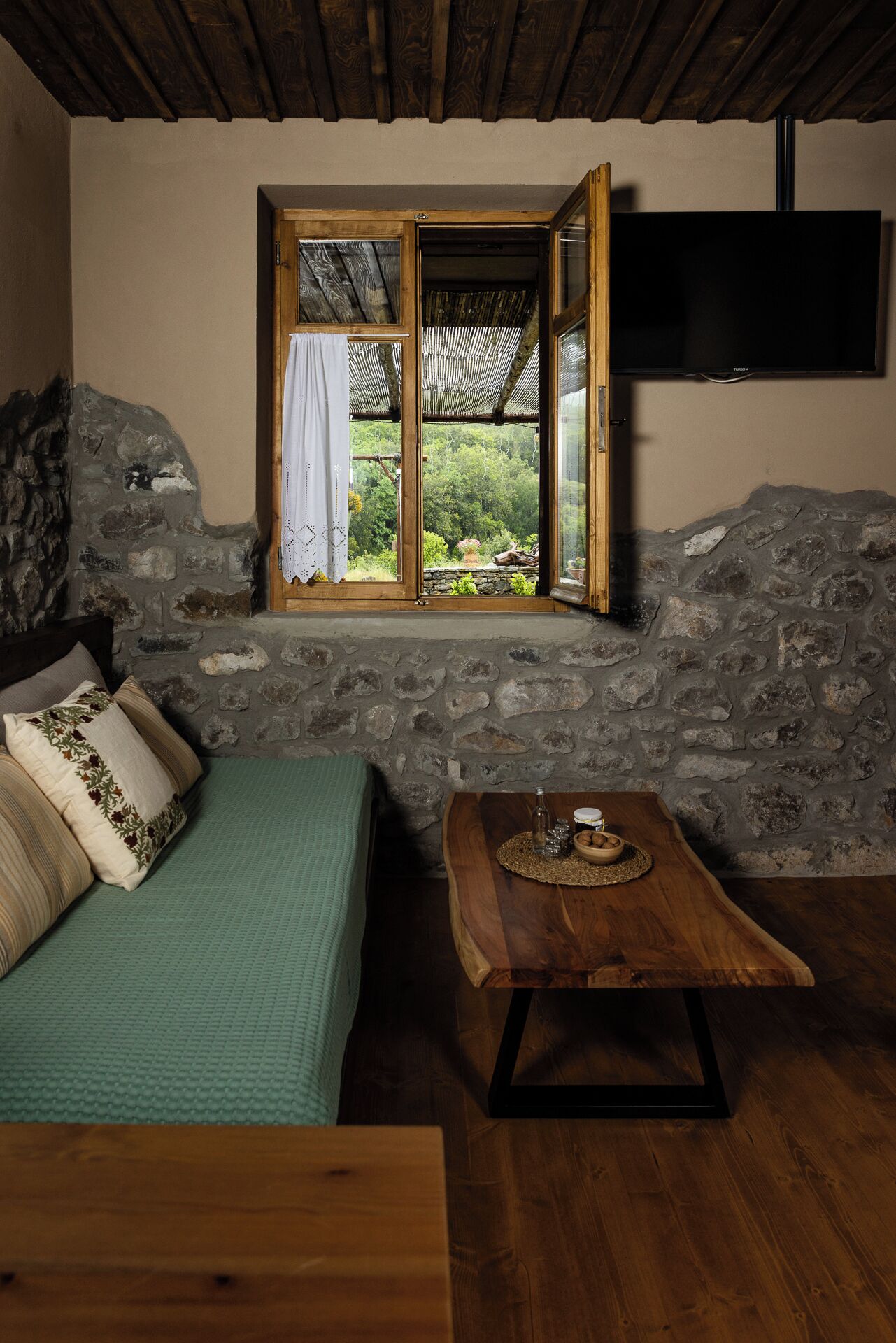 A rustic room with a stone wall, a wooden bed, a table with a bottle and snacks, and a TV mounted on the wall.
