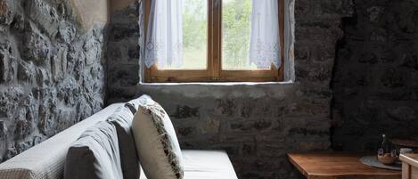 A rustic room with a stone wall, a wooden ceiling, a bed with pillows, a wooden table, and a window with curtains.