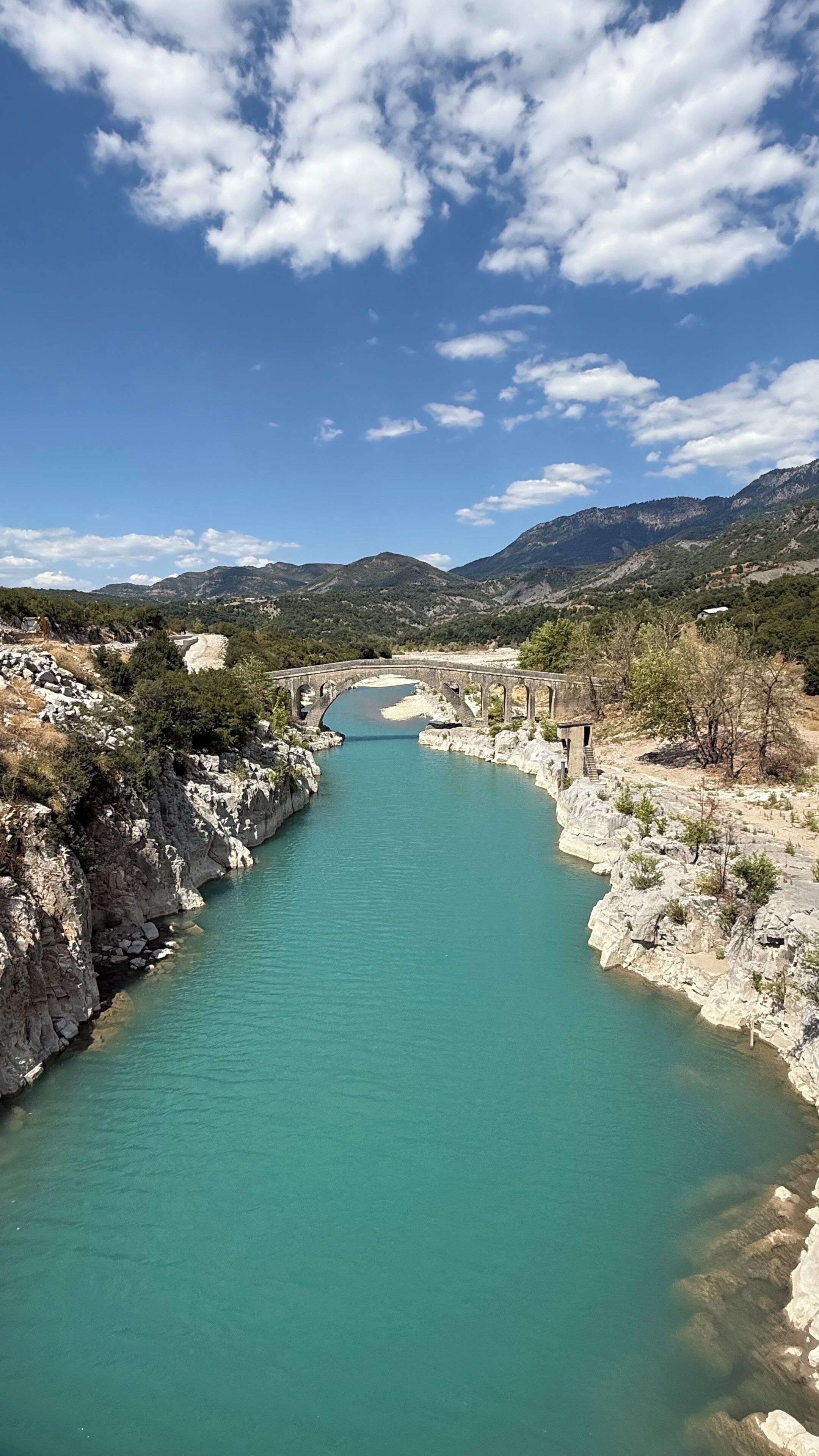 A river with a bridge, rocky banks, and distant mountains under a clear sky.