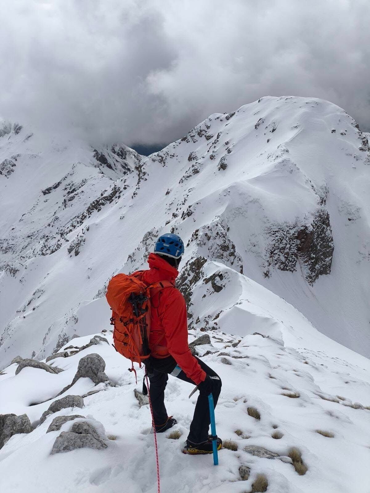 A person in red and blue gear with a large orange backpack, standing on a snowy mountain slope with rocky outcrops.